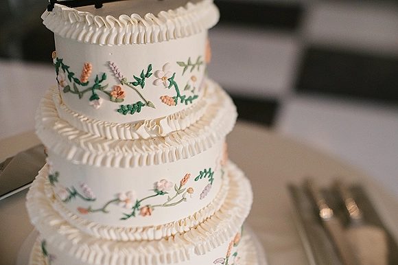 Wedding cake with white buttercream frosting, tiered ruffle borders and hand-piped floral icing on a stand on a tabletop with forks