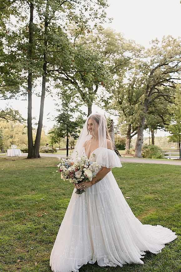 Bridal portrait of a bride holding bouquet in a flowing wedding dress and veil on a park path with trees and greenery behind