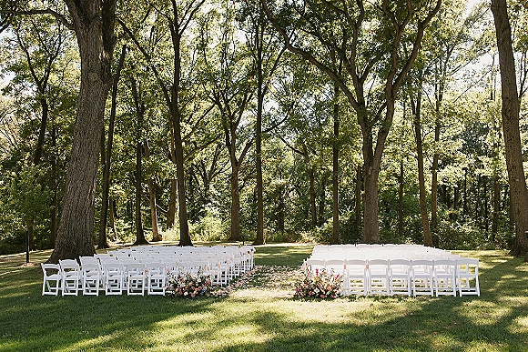 Outdoor ceremony setup with white folding chairs lining a wide forest wedding aisle, scattered petals and flower arrangements on a sunlit lawn