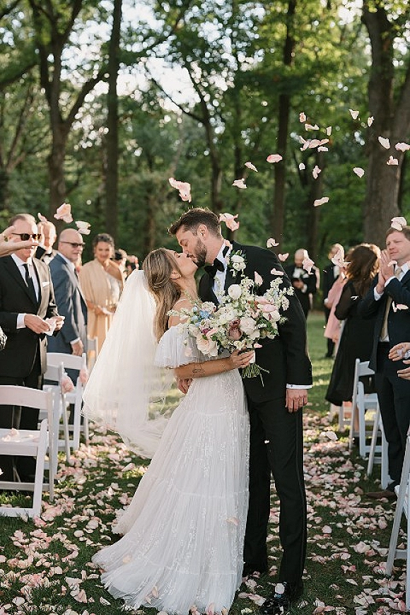 Wedding kiss as newlyweds walk down the aisle with bouquet and veil, rose petals falling among guests under tree-lined lawn ceremony