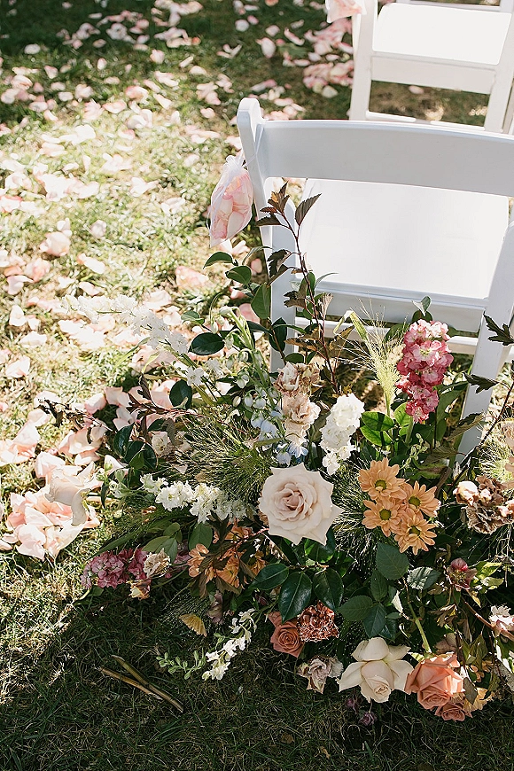 Ceremony aisle flowers lining an outdoor lawn aisle with wedding aisle petals, low rose and mixed bloom arrangements, and white folding chairs
