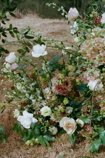 Wedding florals in a ground floral arrangement with blush and white roses, hydrangea, and greenery on pine straw beneath garden trees