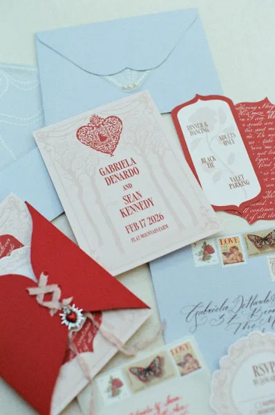 Wedding invitation suite with red and white wedding invitations, calligraphy envelopes, wax seal, stamps, and lace on fabric flat lay