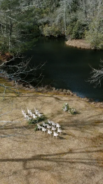 Outdoor ceremony setup with white chairs in a semicircle and floral ground arrangements by a pond, framed by trees and lawn