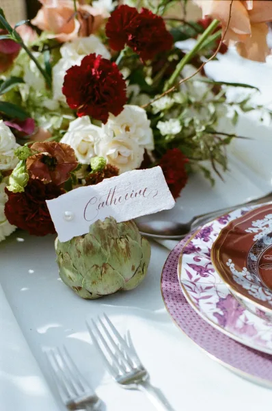 Reception tablescape with wedding place setting, calligraphy place cards in artichoke holders, patterned china, and floral centerpiece on linen tablecloth