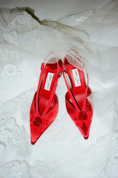 Bridal shoes in red wedding shoes style, red satin heels with heart details and ankle straps resting on lace dress and veil on white bedding