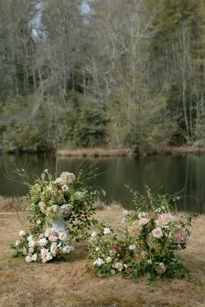 Wedding ceremony flowers in blush and white roses and hydrangeas with greenery, a white pedestal urn, set by a lakeshore with trees