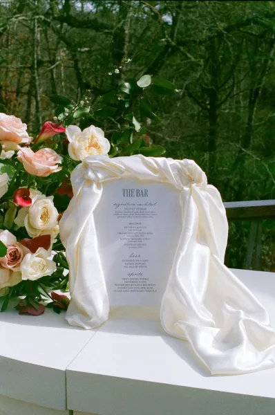 Wedding bar menu sign with modern typography on a white tabletop, framed by satin drape and roses with calla lilies outdoors