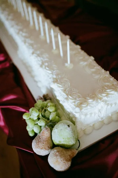 Wedding cake with white sheet cake buttercream rosettes, tall candles, and grapes and pears on pink satin against a dark background