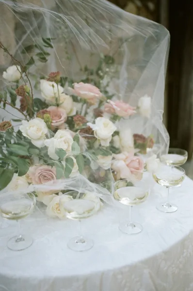 Reception tablescape with a wedding table centerpiece of roses and greenery, bridal veil draped over lace tablecloth and coupe glasses indoors