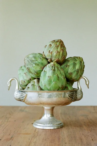 Artichoke centerpiece in a silver swan-handled pedestal bowl on a wooden tabletop against a plain wall, styled as edible wedding decor