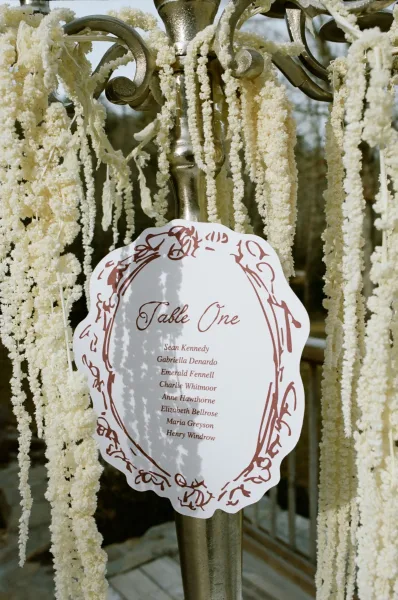 Wedding table sign with calligraphy table number on a candelabra stand, framed by hanging floral garland on an outdoor patio with trees and sky