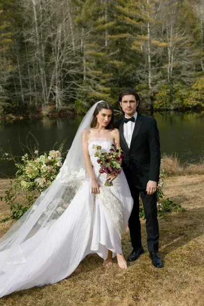Couple portrait of bride in strapless lace gown holding a bouquet beside groom in black tuxedo, with a long veil by a lakeside forest lawn