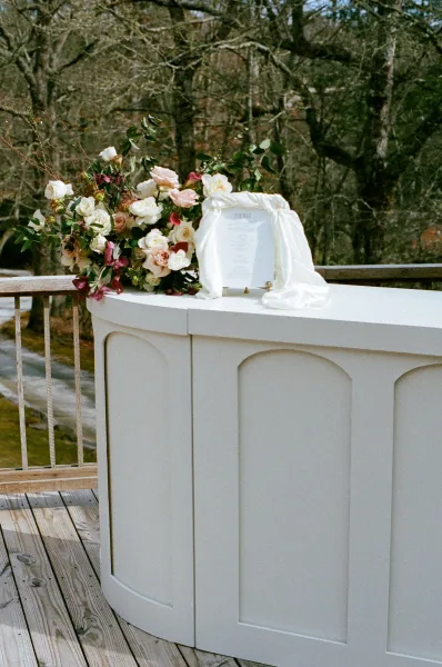 Wedding welcome sign with calligraphy on a brass stand, topped with roses and greenery, set on a wood deck with trees and hillside behind