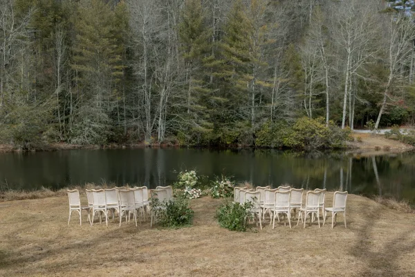 Outdoor ceremony setup with lakefront wedding ceremony seating in a semicircle of white chairs, greenery aisle markers on a grassy lawn by forest trees