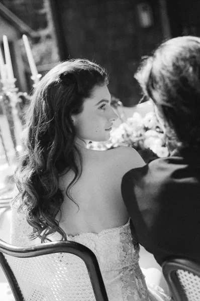 Wedding reception moment with bride looking over shoulder in a strapless lace gown, seated at a candlelit table with floral centerpiece indoors