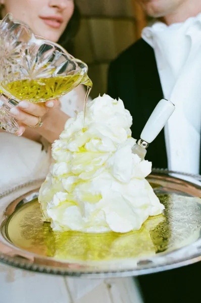 Wedding cake with olive oil being poured from a crystal pitcher onto a white ruffled tier on an antique silver tray indoors