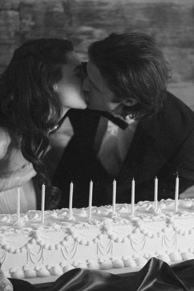 Wedding cake cutting as bride and groom kiss behind a tiered white cake with unlit candles and satin ribbon against stone wall