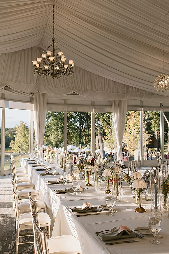 Reception tablescape with a long head table setup, white linens, floral bud vases, taper candles, and gold lamps under a clear tent with chandeliers