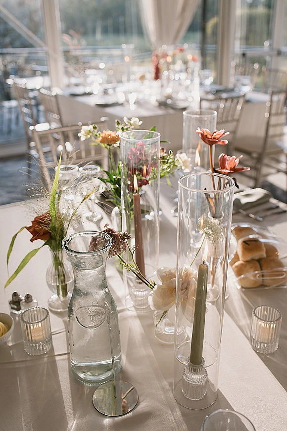 Reception tablescape with wedding table decor featuring taper candles, bud vases, and glass hurricanes on a sunlit patio by glass walls
