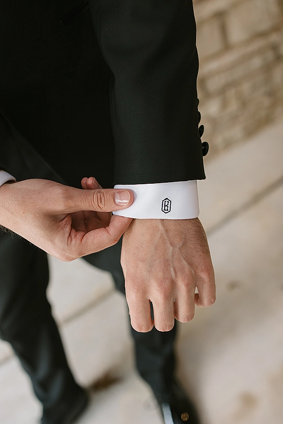 Groom cuff detail showing monogram shirt cuff with embroidered initials, white dress shirt and black suit jacket beside a stone wall walkway