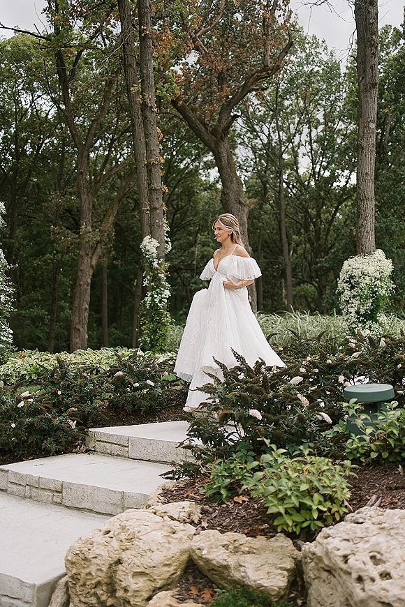 Bridal portrait of a bride holding her full skirt off the shoulder wedding dress on stone steps framed by floral columns and woodland trees