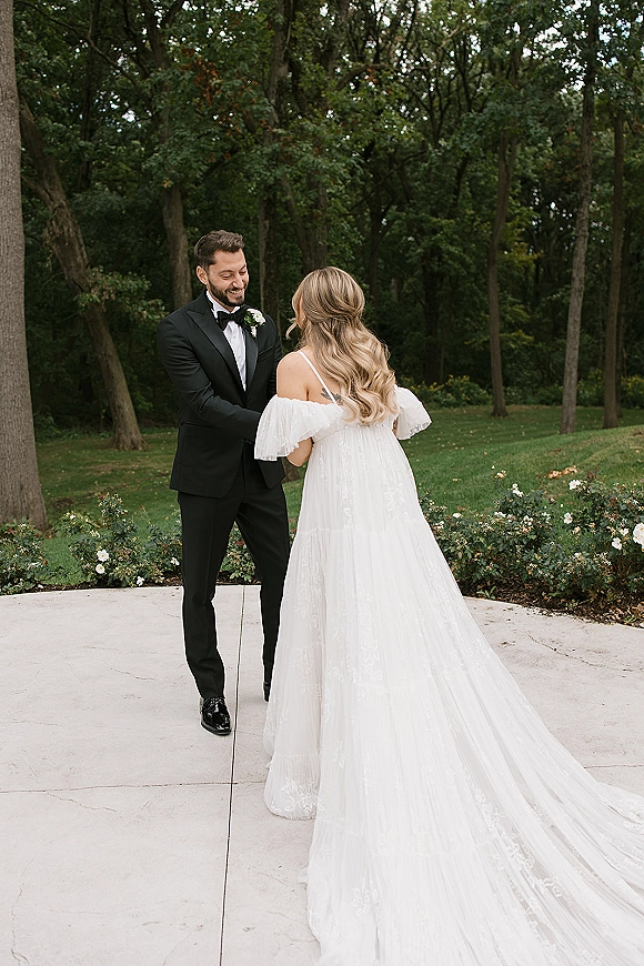 Wedding first look as the groom reaction first look unfolds, bride in off-the-shoulder gown with train holding hands on a garden lawn by rose bushes