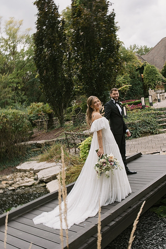 Couple portrait of bride and groom walking hand in hand on a wooden bridge by a pond, bride glancing back in off-shoulder gown