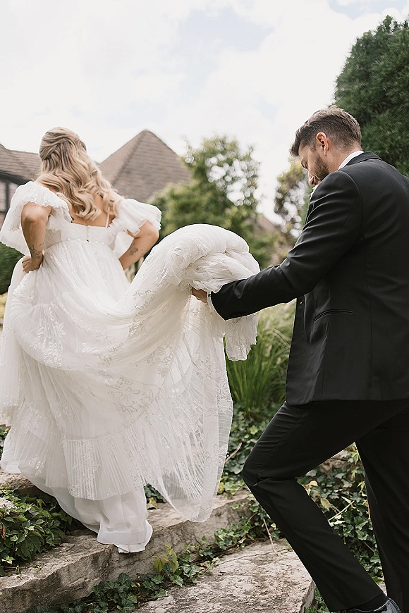 Couple portrait of bride and groom walking up stone steps, groom holding her lace wedding dress train amid garden greenery and ivy