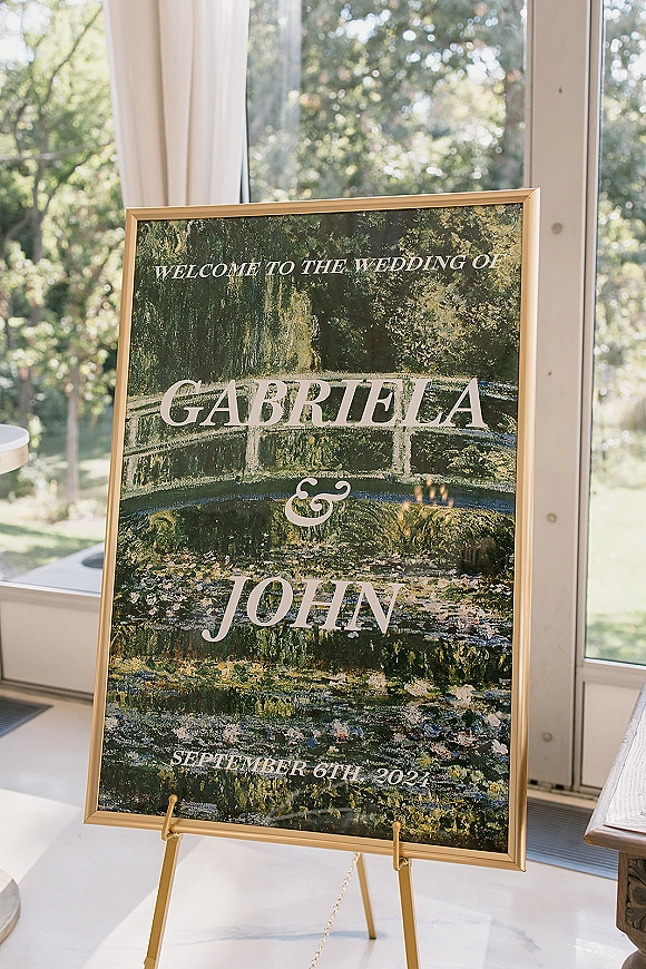 Wedding welcome sign in a gold frame on an easel, printed typography glowing by large windows with white curtains and sunlit greenery outside