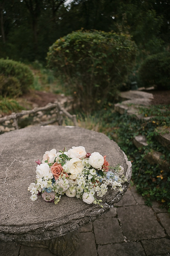 Wedding bouquet of white and peach roses with ranunculus and greenery accents resting on a stone pedestal table in a garden patio setting