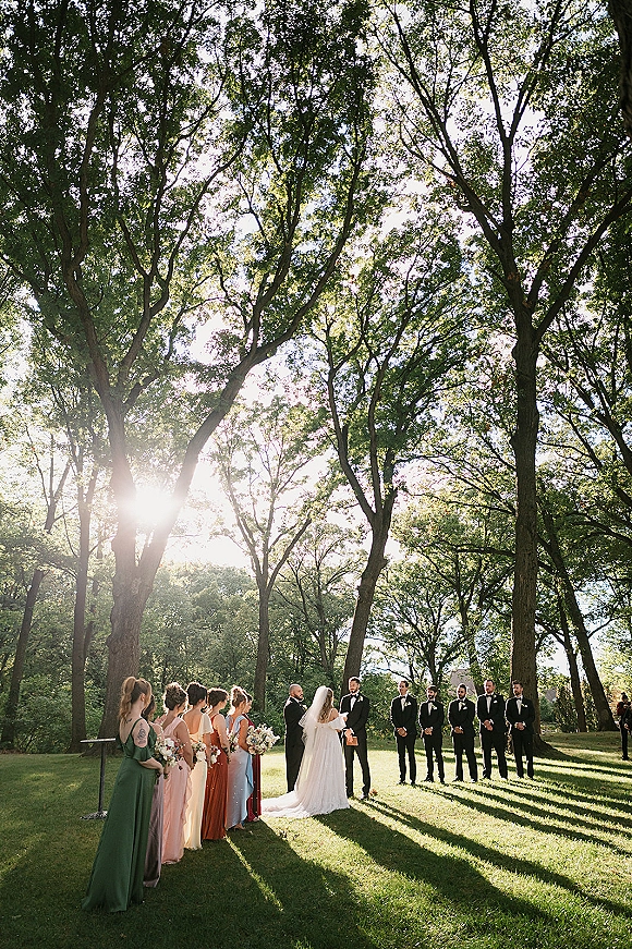 Outdoor wedding ceremony wide shot with bride in long veil and groom at the altar, bridesmaids with bouquets and groomsmen in tuxedos on a sunlit lawn under trees