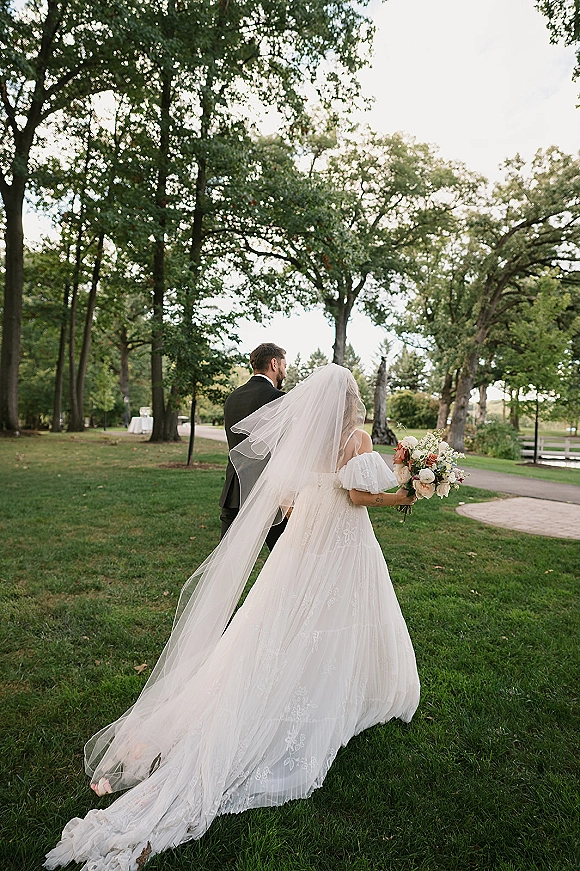 Couple portrait of bride and groom walking away, her long veil train and rose bouquet flowing along a lakeside park path