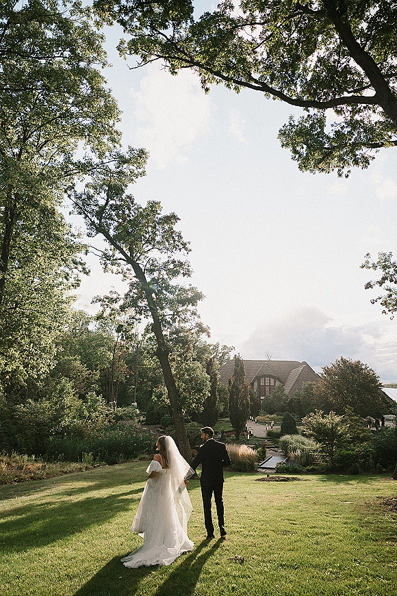 Couple portrait of newlyweds holding hands walking away, bride in veil and gown with train, groom in black tux on garden lawn