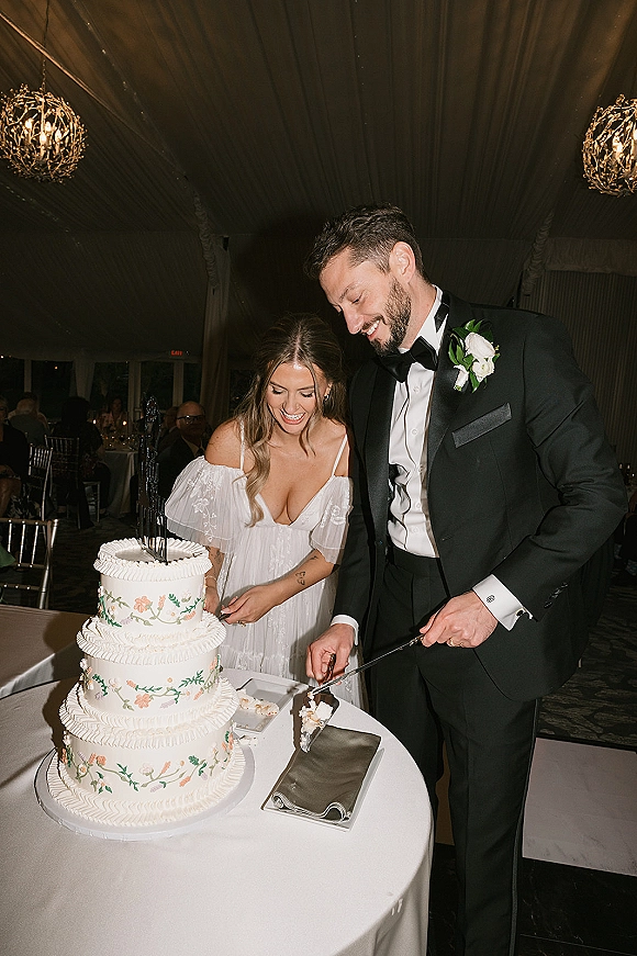 Wedding cake cutting as bride in off-shoulder gown and groom in black tux slice a three-tier hand-painted floral cake under tent chandeliers