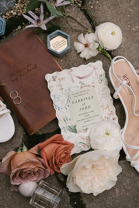 Wedding invitation suite with illustrated wedding invitation, vow book, gold rings and sandals, surrounded by pastel flowers on stone pavement