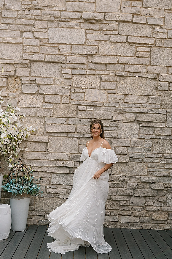 Bridal portrait of a bride standing by a stone wall, holding her lace off-the-shoulder gown with a long train and tiered skirt