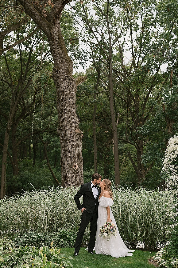 Wedding kiss portrait of bride and groom kissing, bride holding bouquet in off-shoulder gown beside groom in tux, amid woodland trees and tall grass