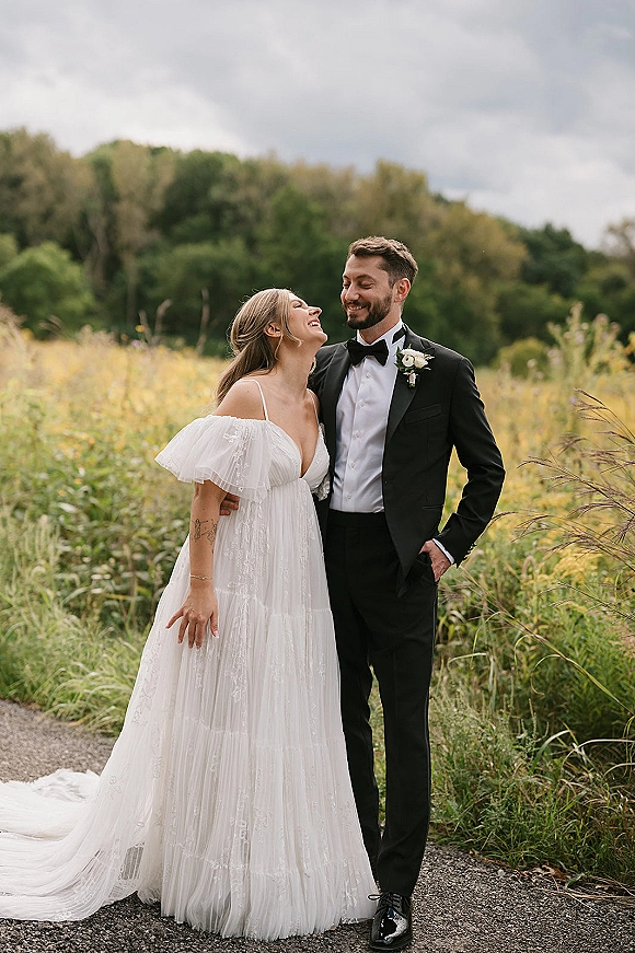 Couple portrait of bride and groom laughing as he hugs her on a wildflower field path, her off-shoulder gown train flowing under cloudy sky