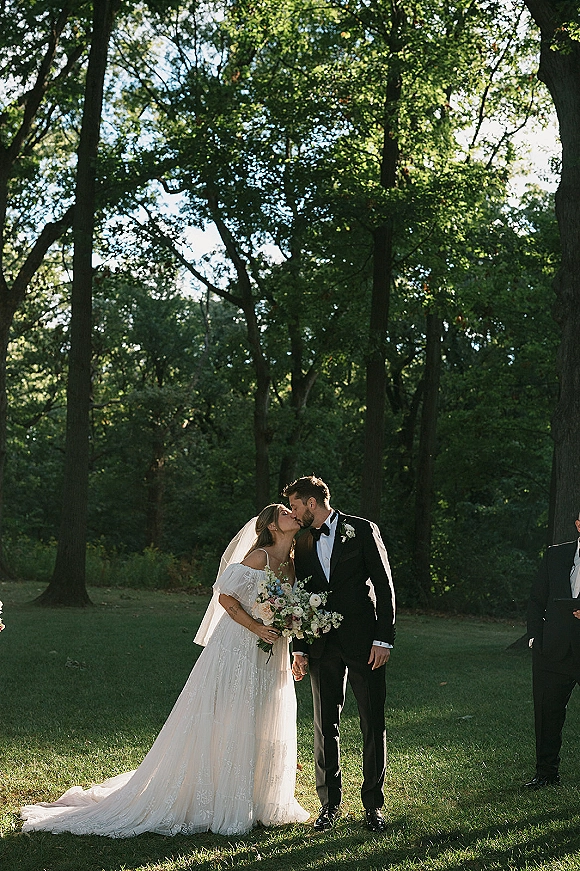 Wedding kiss portrait of bride and groom kissing on a forest lawn, holding hands as she holds a bouquet with veil flowing