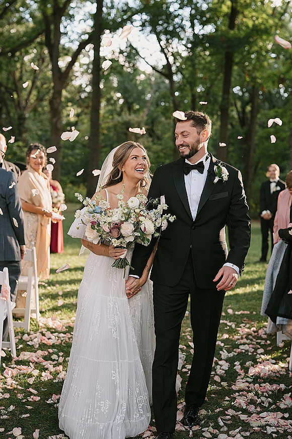 Wedding recessional as bride and groom walk the aisle hand in hand, bouquet and veil trailing through tossed petals in a garden ceremony