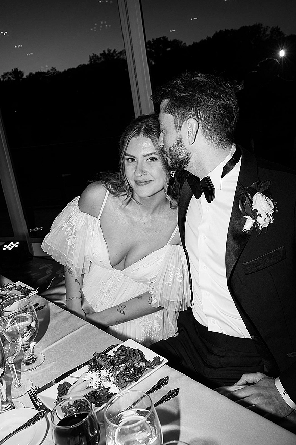 Couple portrait in a black and white wedding portrait style, groom kisses bride’s cheek at sweetheart table by windows at night