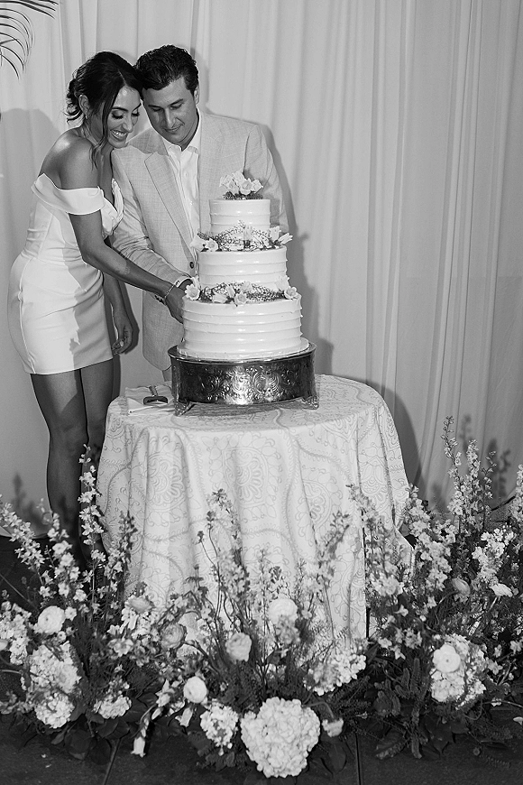Cake cutting moment as bride and groom slice a three tier wedding cake with fresh flowers at a draped reception backdrop