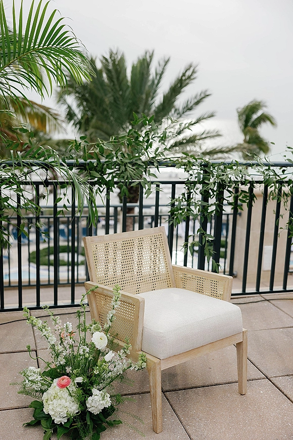 Wedding ceremony chair with cane back and white cushion, adorned with white and pink florals and greenery on an ocean-view balcony