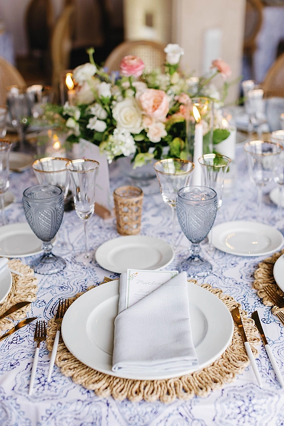 Reception tablescape with wedding table setting, rose floral centerpiece, taper and votive candles, blue goblets, gold flatware, blurred guests behind