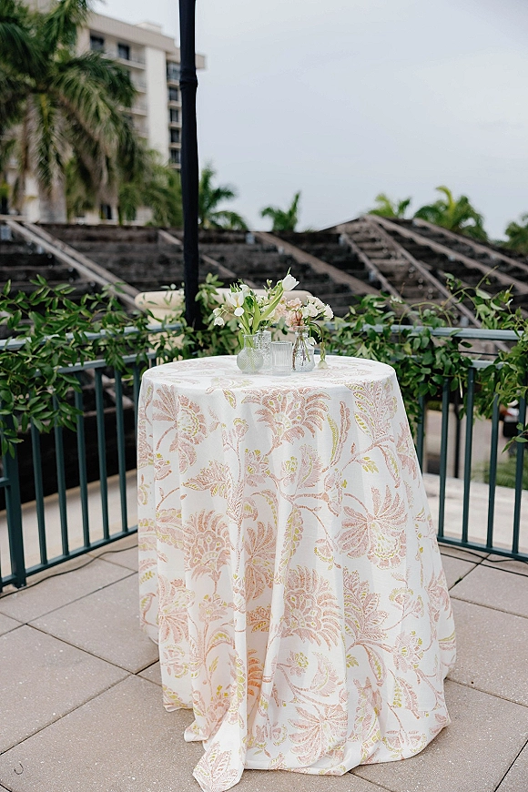 Cocktail table decor with patterned linen, bud vases of white flowers and greenery, glassware and a candle on a palm-lined terrace