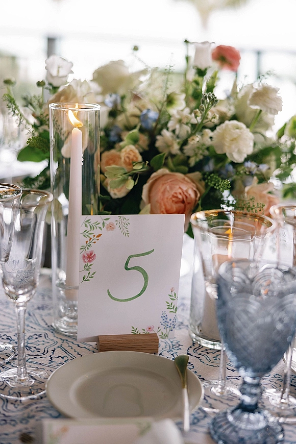Wedding tablescape with wedding table centerpiece of white and blush flowers, taper candle in hurricane, blue goblets, and table number card in bright room