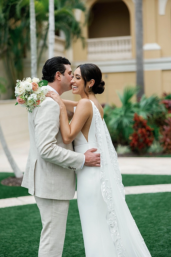 Wedding couple portrait of bride and groom embrace, her veil and bouquet visible, in a palm-lined courtyard with arched building backdrop