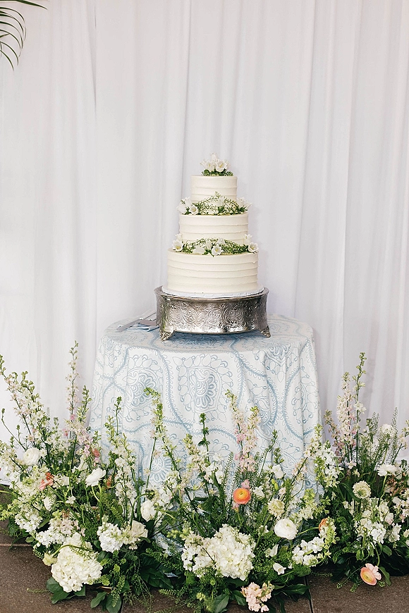 Wedding cake with greenery garland, three-tier white buttercream with floral topper on a silver stand before white drapery backdrop