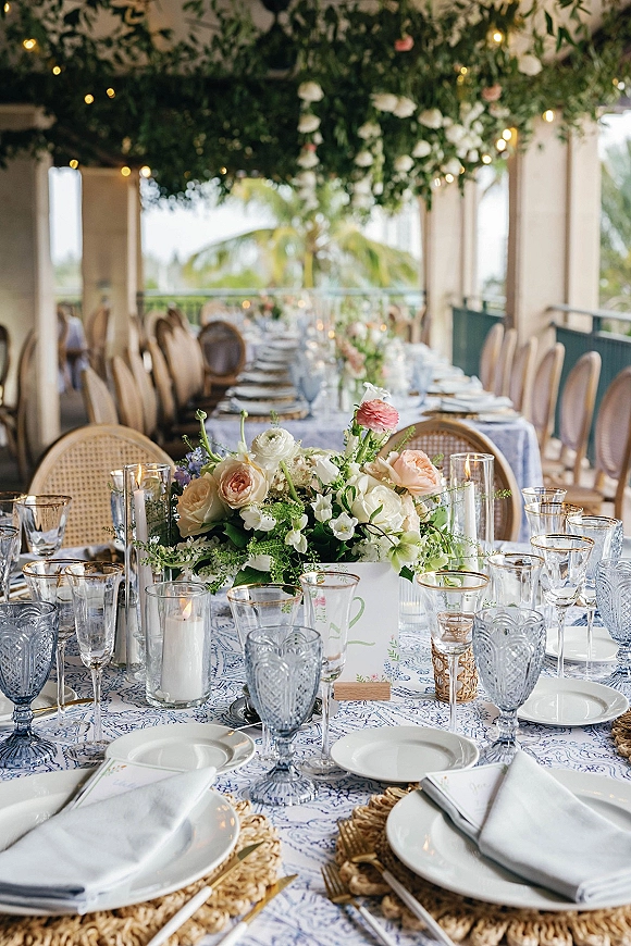 Reception tablescape with blue and white wedding tablescape details, floral centerpieces, taper candles, and string lights on a veranda with palms beyond